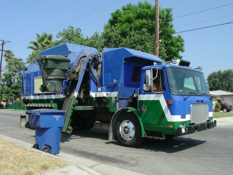 A large blue and green waste collection truck parked on a residential street with a clear blue sky overhead. The vehicle features a front cab with a silver grille and windows, and a rear compactor section painted in a darker shade of blue. Visible on the side are hydraulic arms and mechanisms used for lifting and compacting rubbish, with a green waste bin positioned at the rear left side of the truck. The setting includes a paved driveway or street lined with grass and mature trees in the background, contributing to a suburban environment. The scene is well-lit by natural sunlight, highlighting the clean, metallic textures of the truck and the fresh, green foliage nearby. This image exemplifies professional rubbish collection services by Commercial Waste Kingston, tailored for efficient waste removal within Kingston or surrounding areas, supporting local waste management needs.