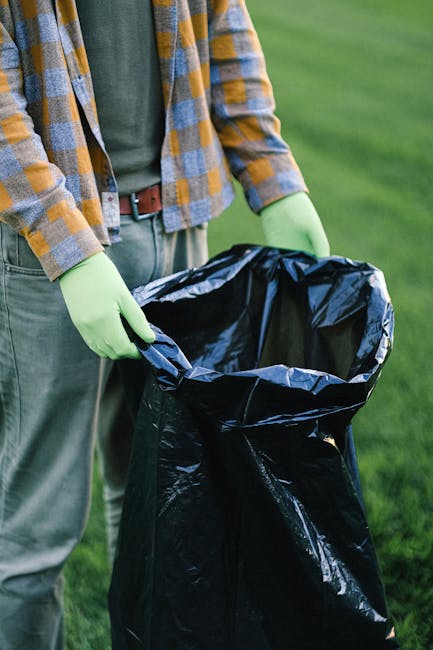 In the foreground, there is a collection of grey plastic rubbish bags with a slightly textured surface, all neatly stacked on a paved driveway. Behind the bags, a large grey skip bin made of metal wit