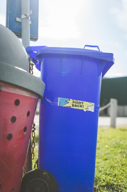 A person's hand holds a green plastic rubbish bag by its twisted knot at the top, with the bag appearing semi-transparent and bulging with contents. The bag is situated in the foreground against a pla