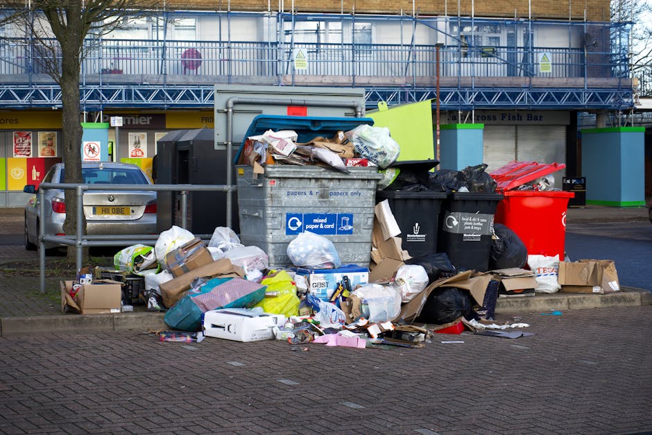 A collection of various types of waste and rubbish placed on a paved outdoor surface, including large cardboard boxes, plastic containers, and discarded packaging materials. The boxes are visibly crum